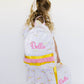 A young girl with blonde hair in a ponytail is shown from behind, wearing a white backpack and matching dress set with pastel rainbow sprinkles and pink 'Bella' script embroidery. She is holding a matching lunch bag against a plain white wall.