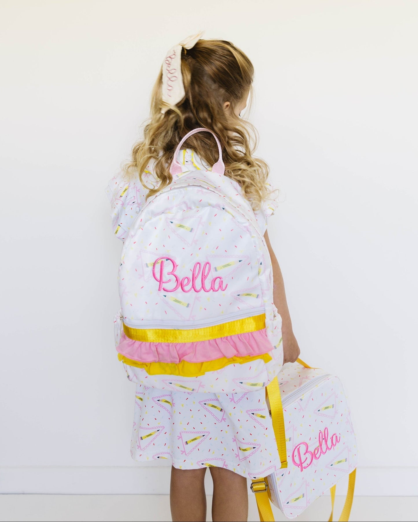 A young girl with blonde hair in a ponytail is shown from behind, wearing a white backpack and matching dress set with pastel rainbow sprinkles and pink 'Bella' script embroidery. She is holding a matching lunch bag against a plain white wall.
