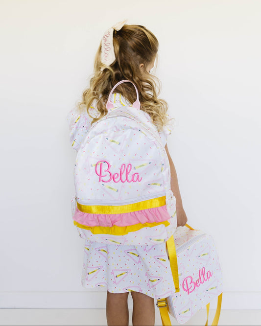 A young girl with blonde hair in a ponytail is shown from behind, wearing a white backpack and matching dress set with pastel rainbow sprinkles and pink 'Bella' script embroidery. She is holding a matching lunch bag against a plain white wall.